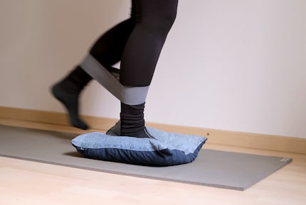 Währenddem das eine Bein auf dem Kissen steht, wird das andere im Halbkreis von vorne nach hinten gegen den Widerstand des Fitnessbands bewegt.